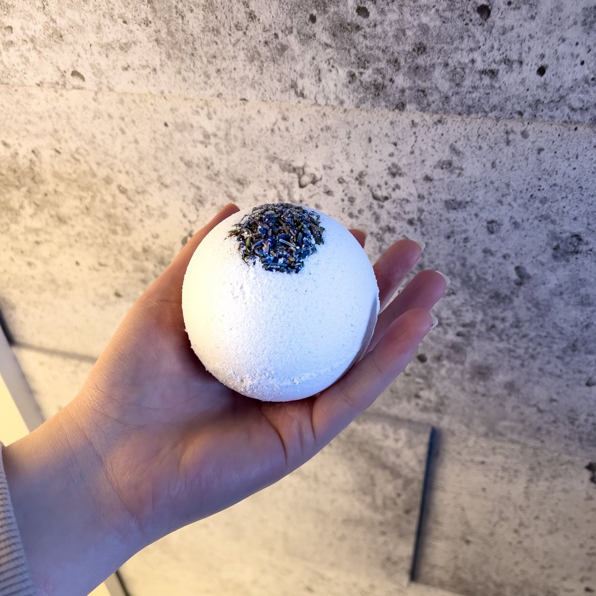 Une bombe de bain blanche soupoudrée de lavande dans une main sur un fond gris